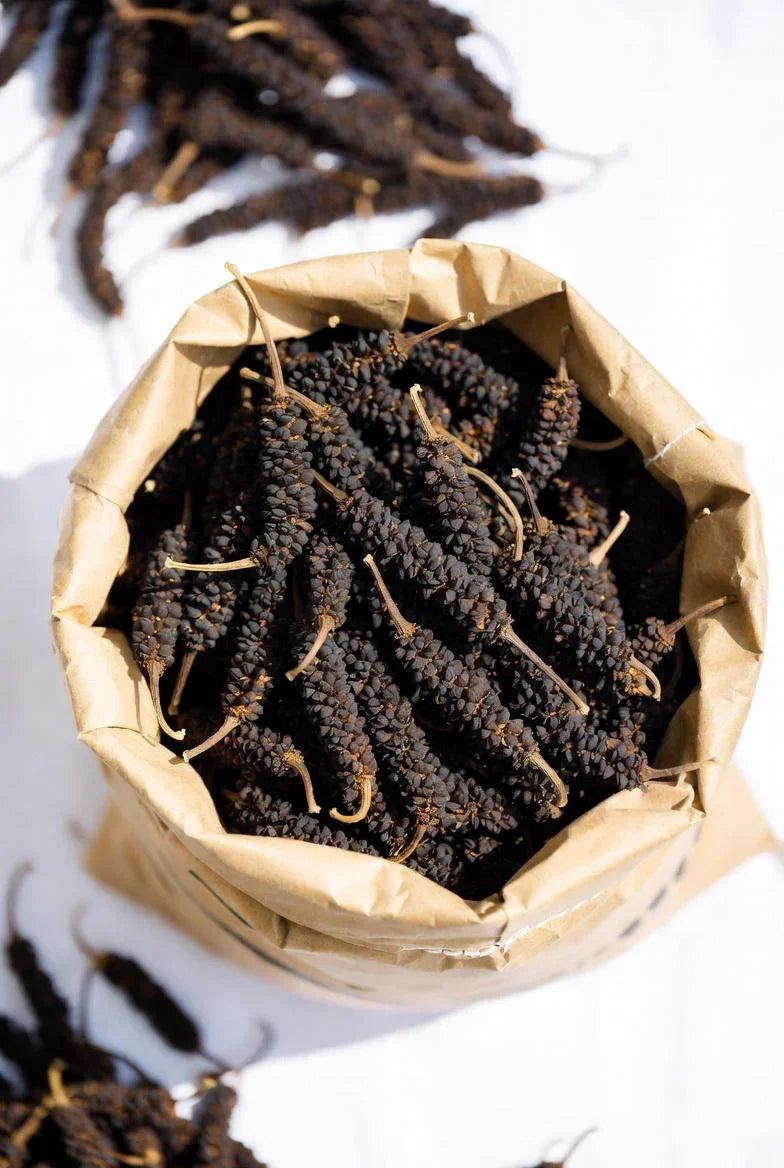 Close-up of dried long black peppercorns in a brown paper bag on a white surface