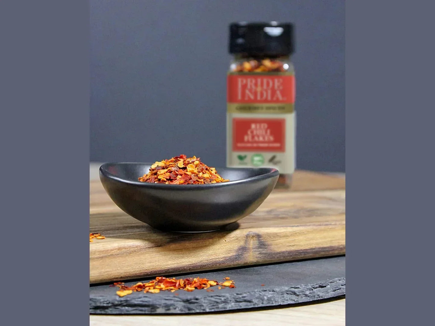 Bowl of red chili flakes with spice jar on wooden board, seasoning closeup