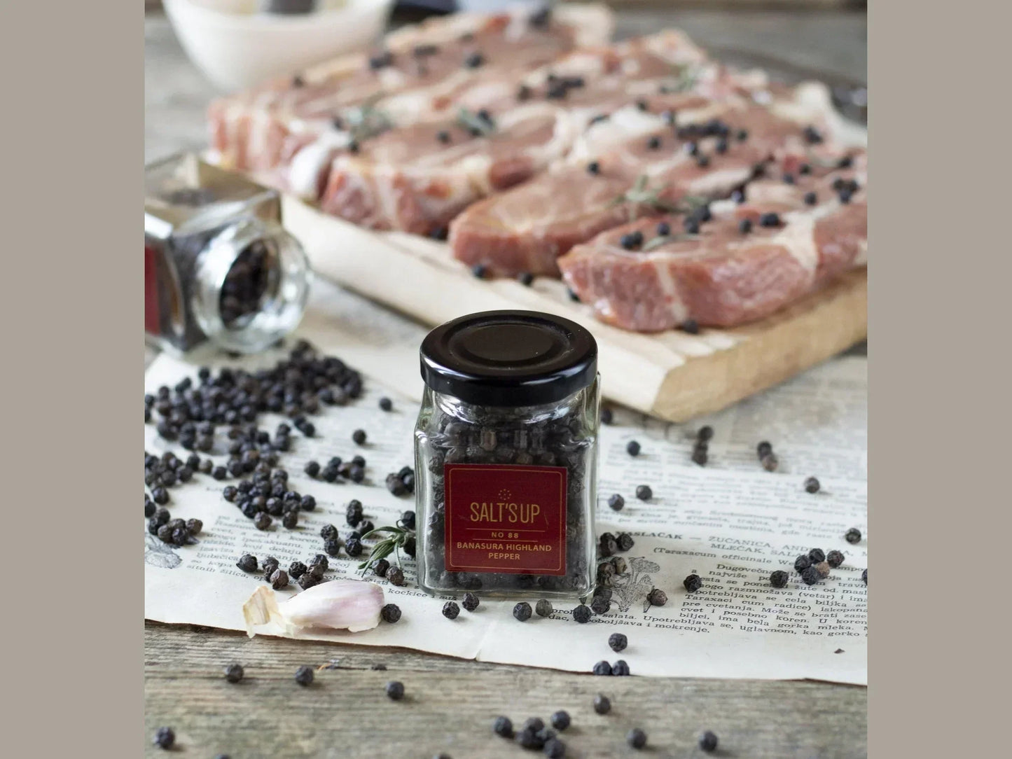 Jar of Banasura Highland black pepper, peppercorns, raw meat, and garlic on rustic table