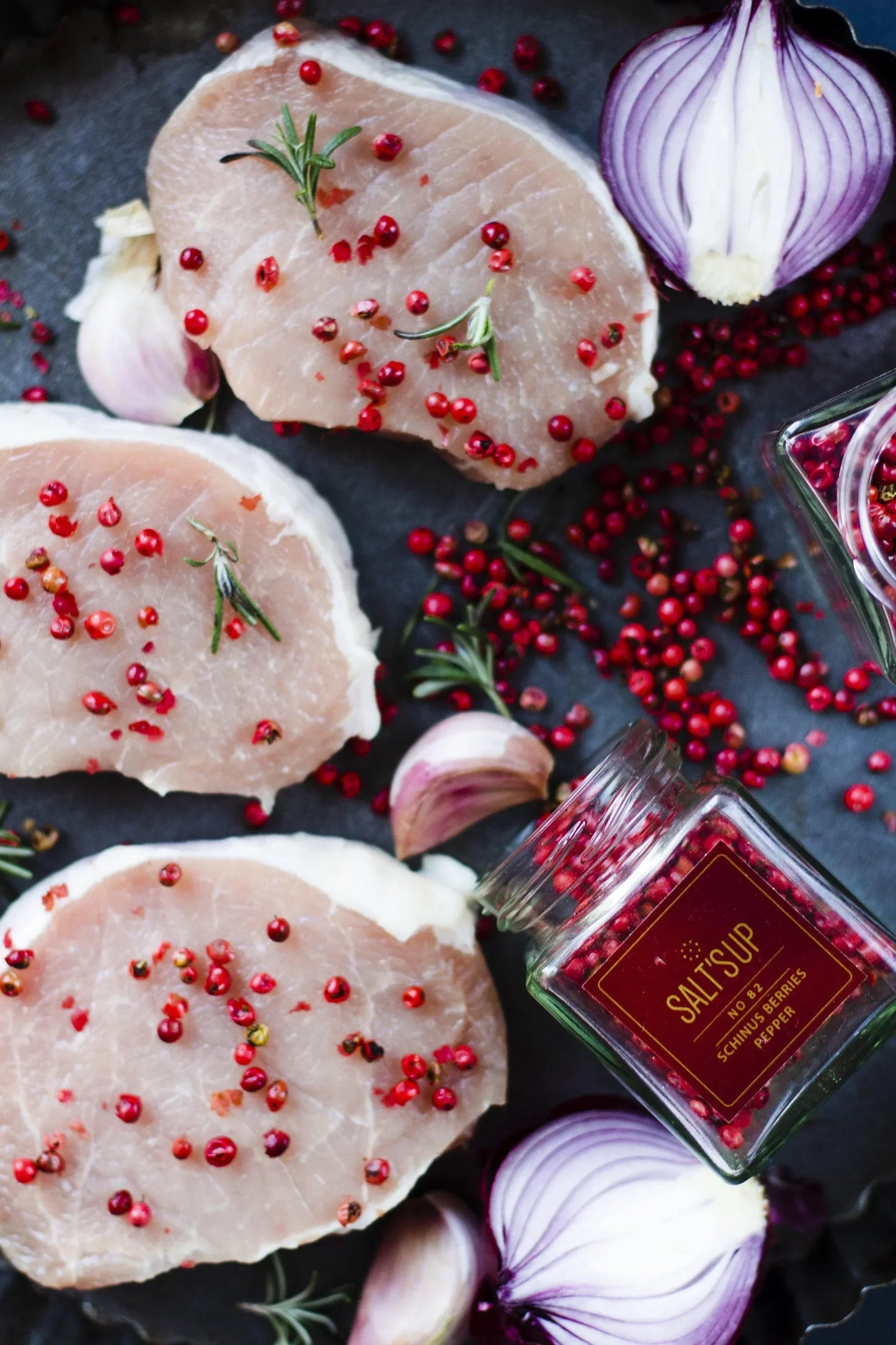 Raw pork steaks topped with schinus berries pepper, fresh rosemary, red onion, and garlic cloves on a dark surface, with a SALTSUP spice jar featured—Spice n Slice Market gourmet ingredients display.