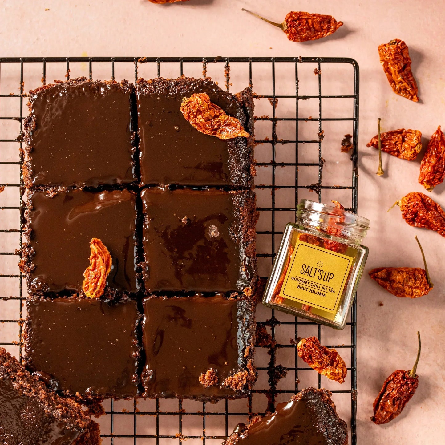 Chocolate brownies with Bhut Jolokia dried chili, gourmet chili jar, and whole dried chilies on a cooling rack.