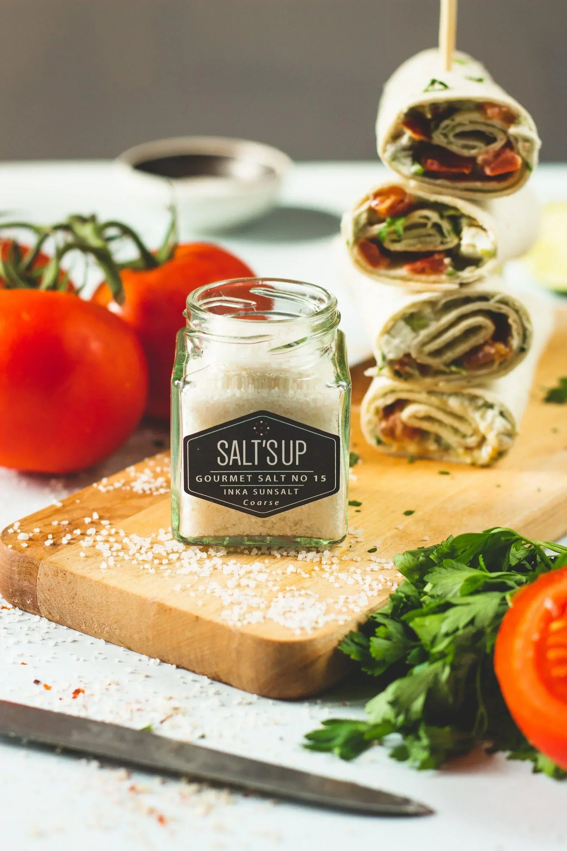Jar of gourmet coarse salt on cutting board with fresh tomatoes, herbs, and vegetable wraps