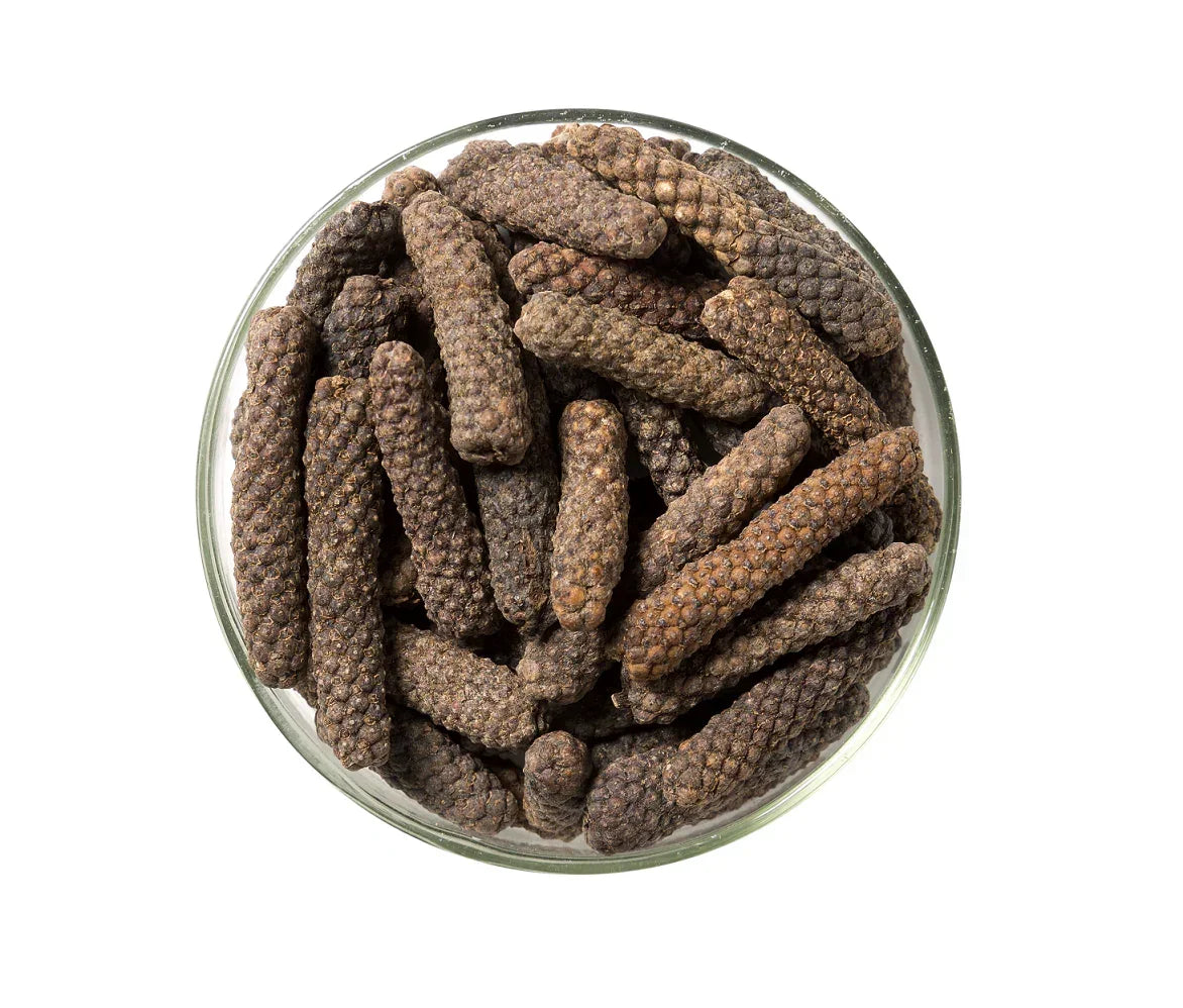 Glass bowl filled with long pepper spice, dark brown textured peppercorns on white background