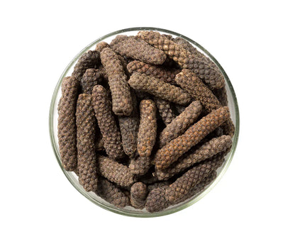 Glass bowl filled with long pepper spice, dark brown textured peppercorns on white background