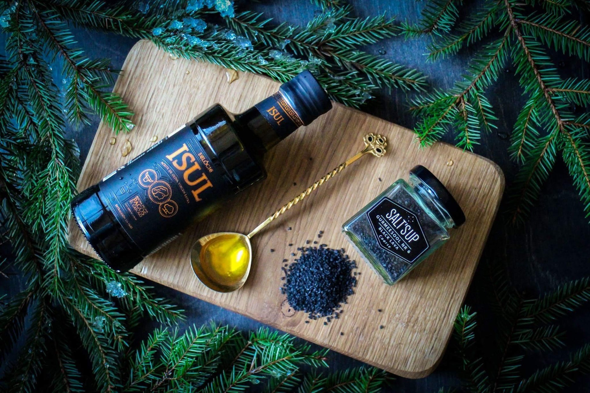Artisan olive oil, black salt, and gold spoon on wooden board with pine branches, gourmet spices
