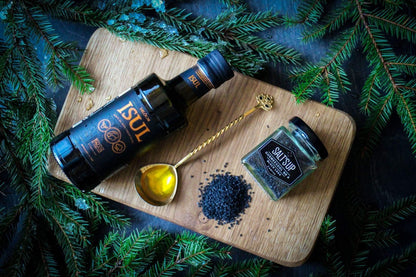 Artisan olive oil, black salt, and gold spoon on wooden board with pine branches, gourmet spices