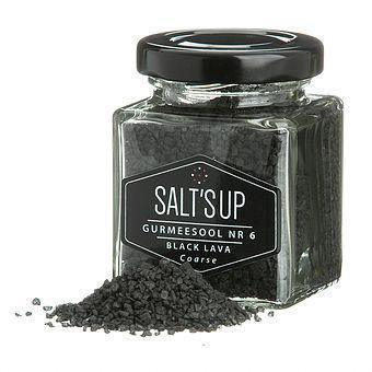 Glass jar of coarse black lava sea salt with scattered crystals on a white background.