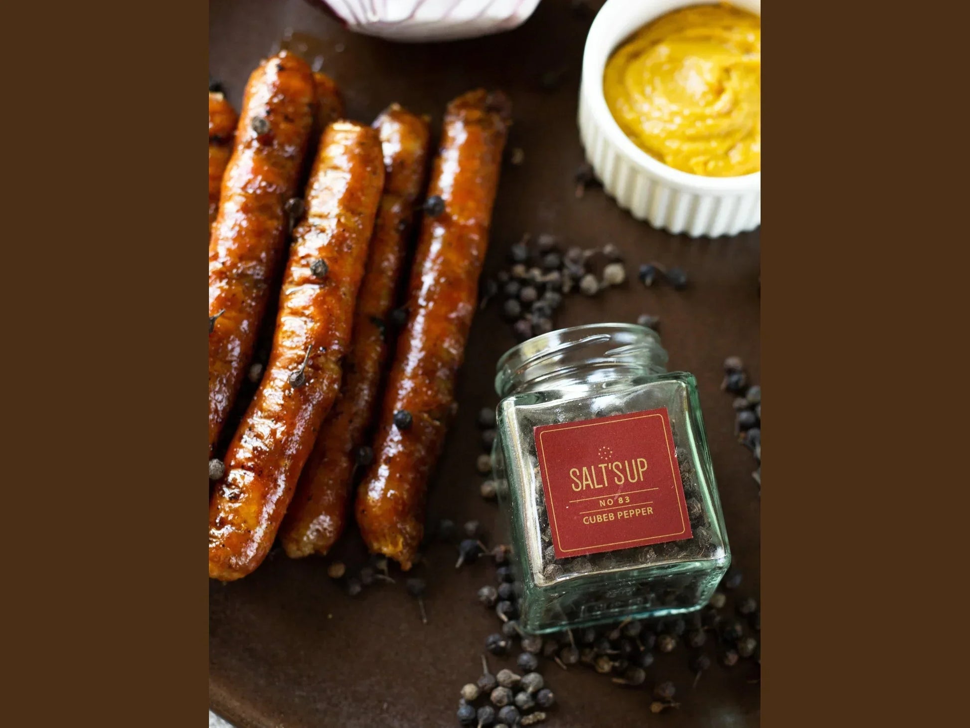Grilled sausages with cubeb pepper, jar of cubeb pepper, and mustard dip on a plate.