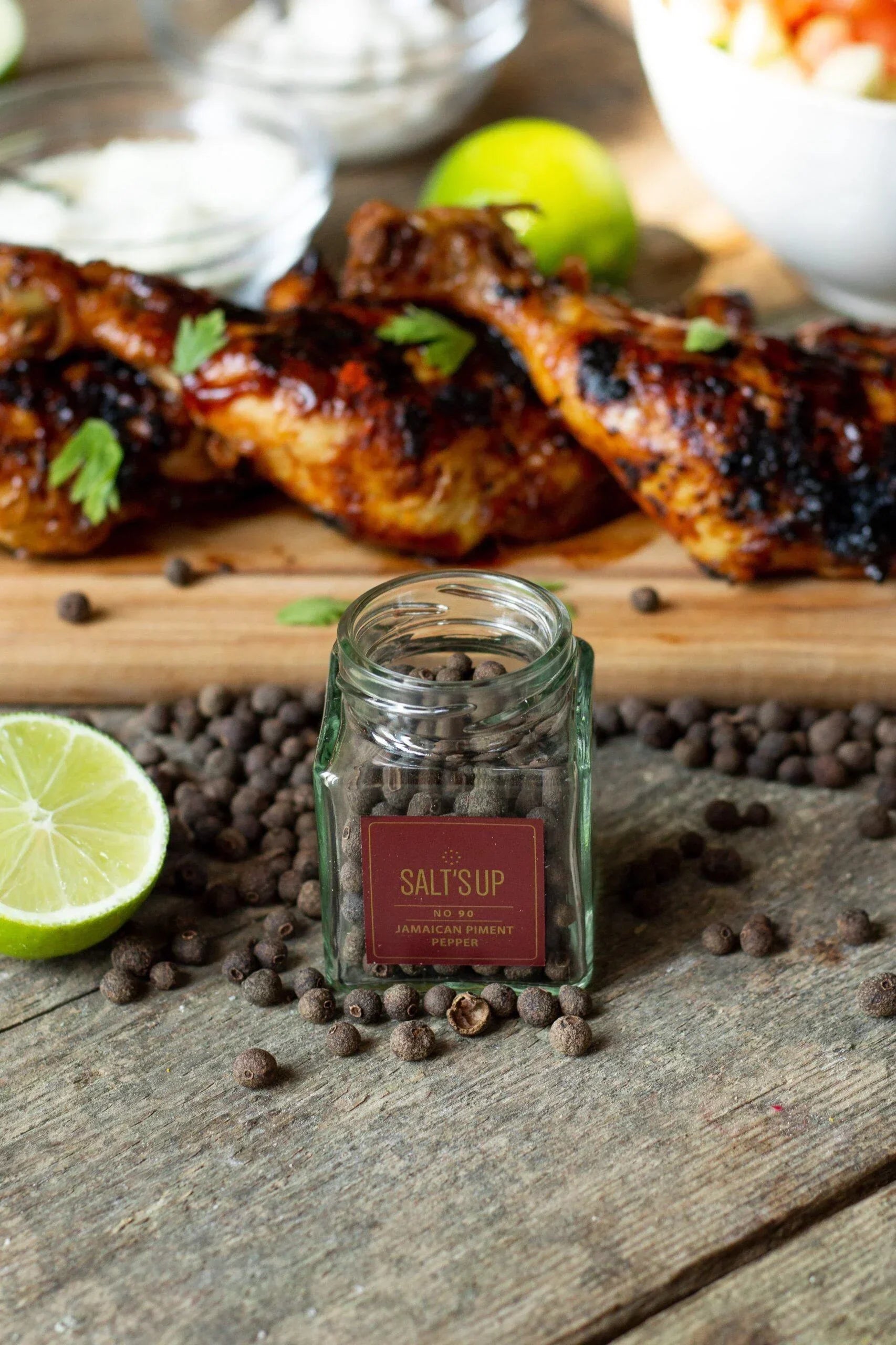 Jamaican piment pepper jar with whole peppercorns, grilled chicken, lime, rustic wood table