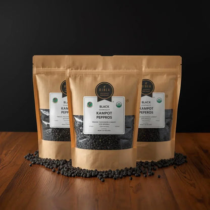 Three brown stand-up pouches of Rinka Black Kampot Peperos on wood surface with black peppercorns scattered around