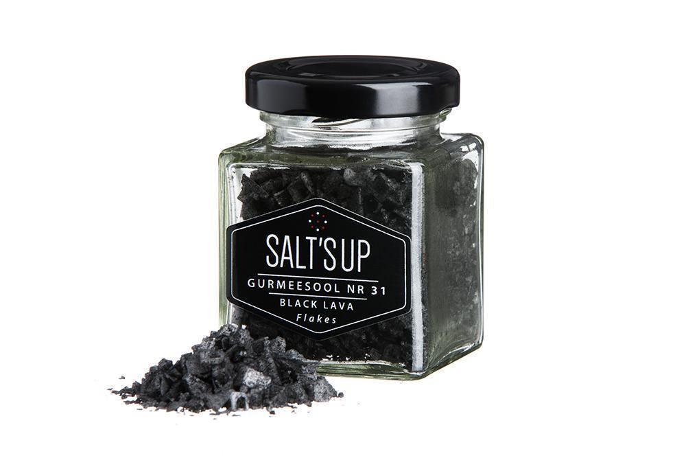 Glass jar of black lava salt flakes with salt flakes spilled in front on white background.
