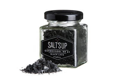 Glass jar of black lava salt flakes with some salt spilled on a white background