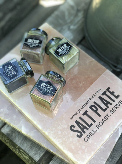 Jars of SALT'SUP gourmet salts on a Himalayan salt plate outdoors, perfect for grilling and roasting.