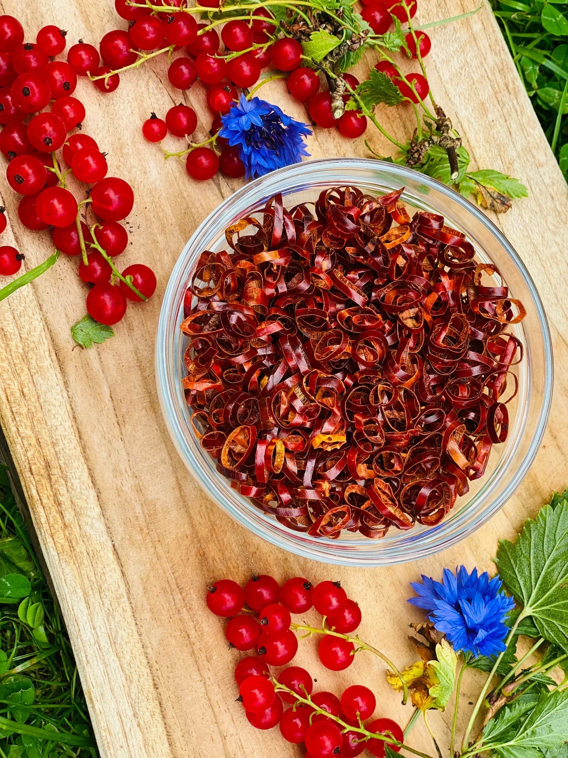 Glass bowl of dried Kampot pepper ribbons on wooden board with fresh red currants and blue flowers