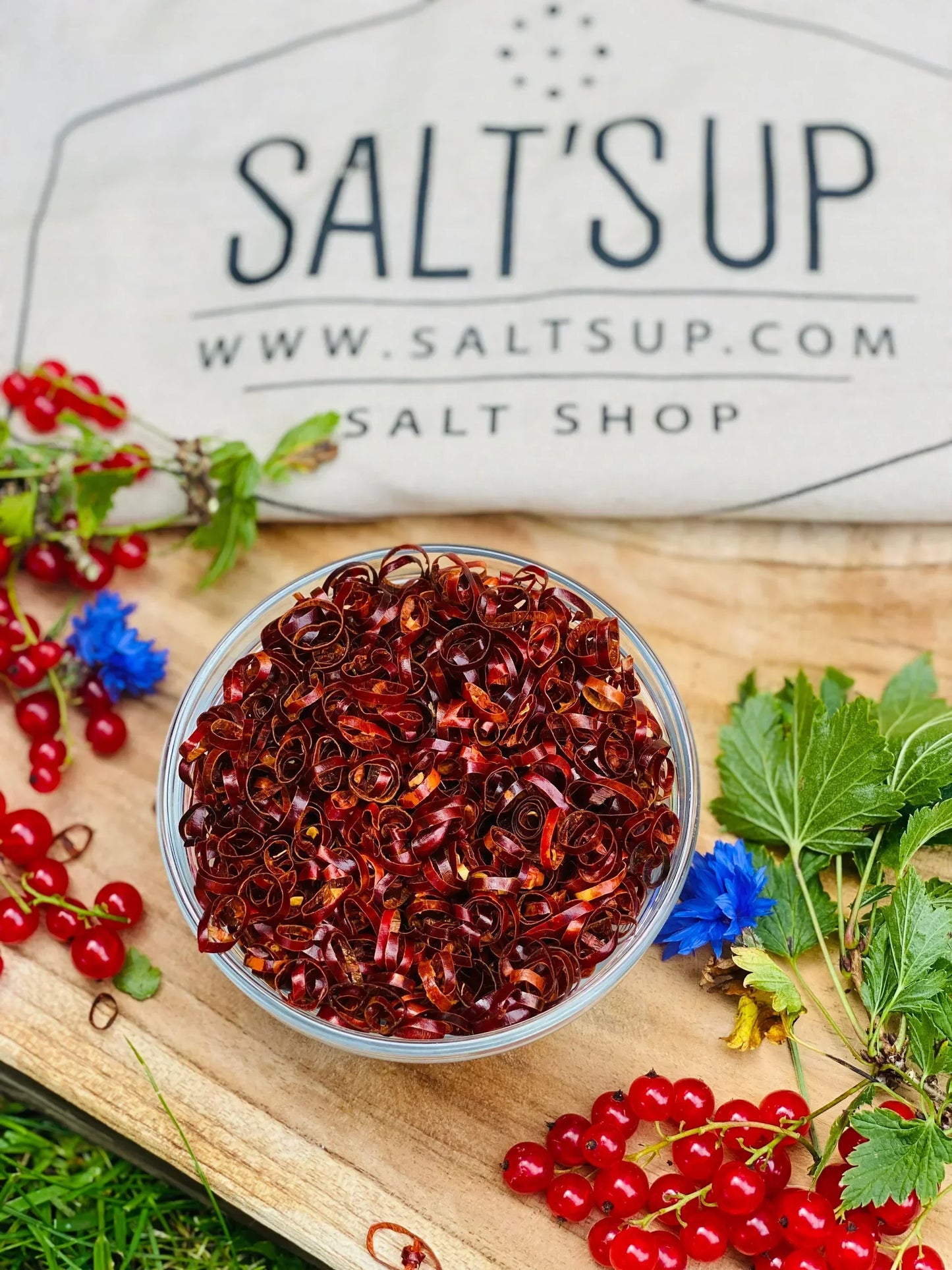 Glass bowl of sliced dried red chili pepper on wooden board with red currants and herbs, Spice n Slice Market specialty.