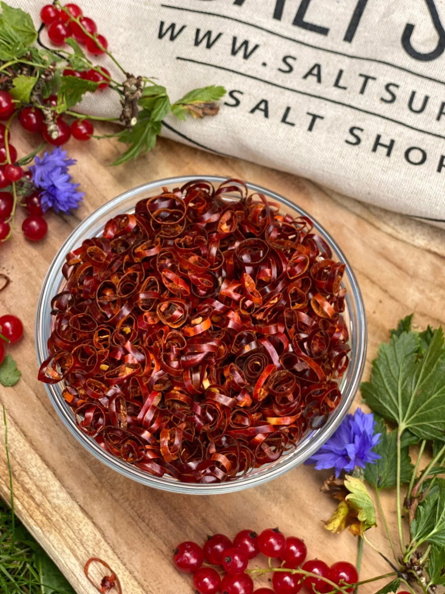 Bowl of dried chili pepper curls on wooden board with currants and flowers