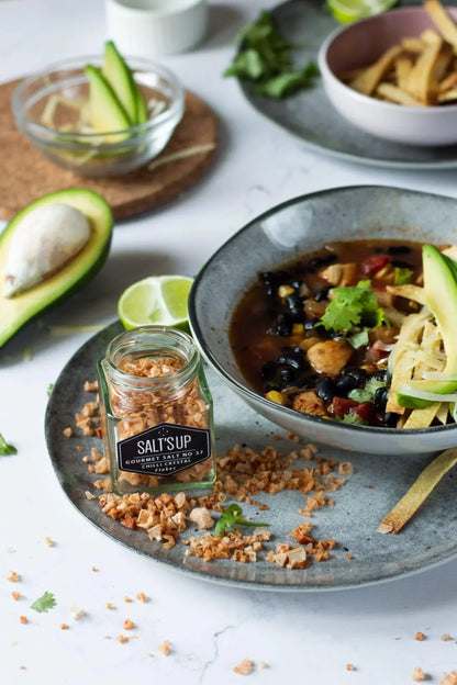 Gourmet chili crystal salt flakes beside avocado, lime, and a bowl of black bean soup on a gray plate.