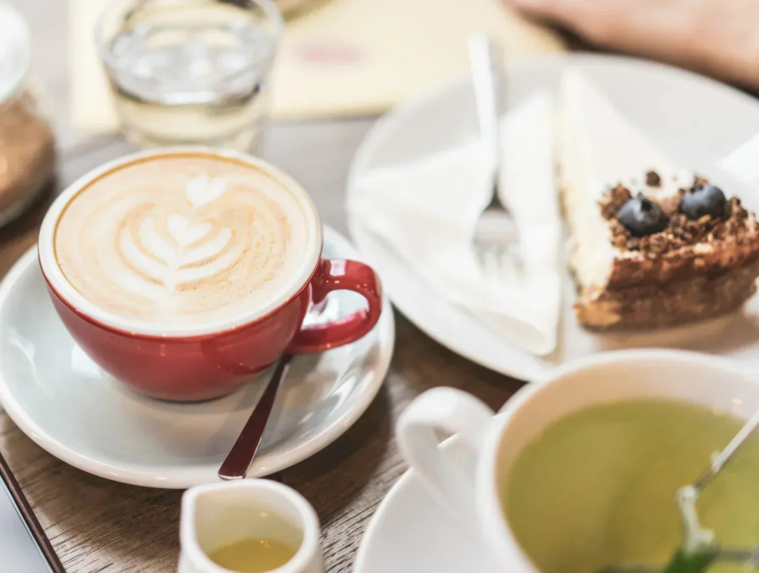 Cappuccino with latte art, cheesecake with blueberries, and green tea on café table