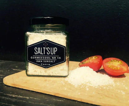 Glass jar of coarse Inka sun salt on wooden board with sliced tomatoes and salt pile