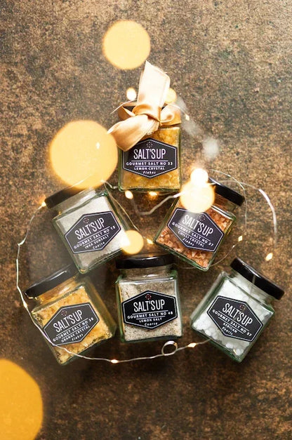 Assorted gourmet salt jars arranged with ribbon and decorative lights on a rustic surface