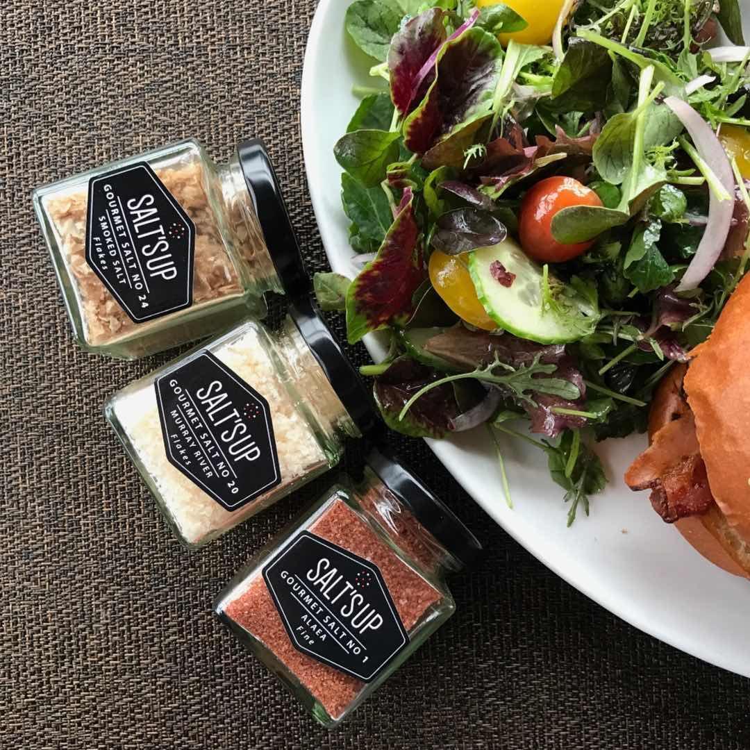 Three jars of gourmet salt next to a fresh salad with mixed greens and vegetables on a plate.