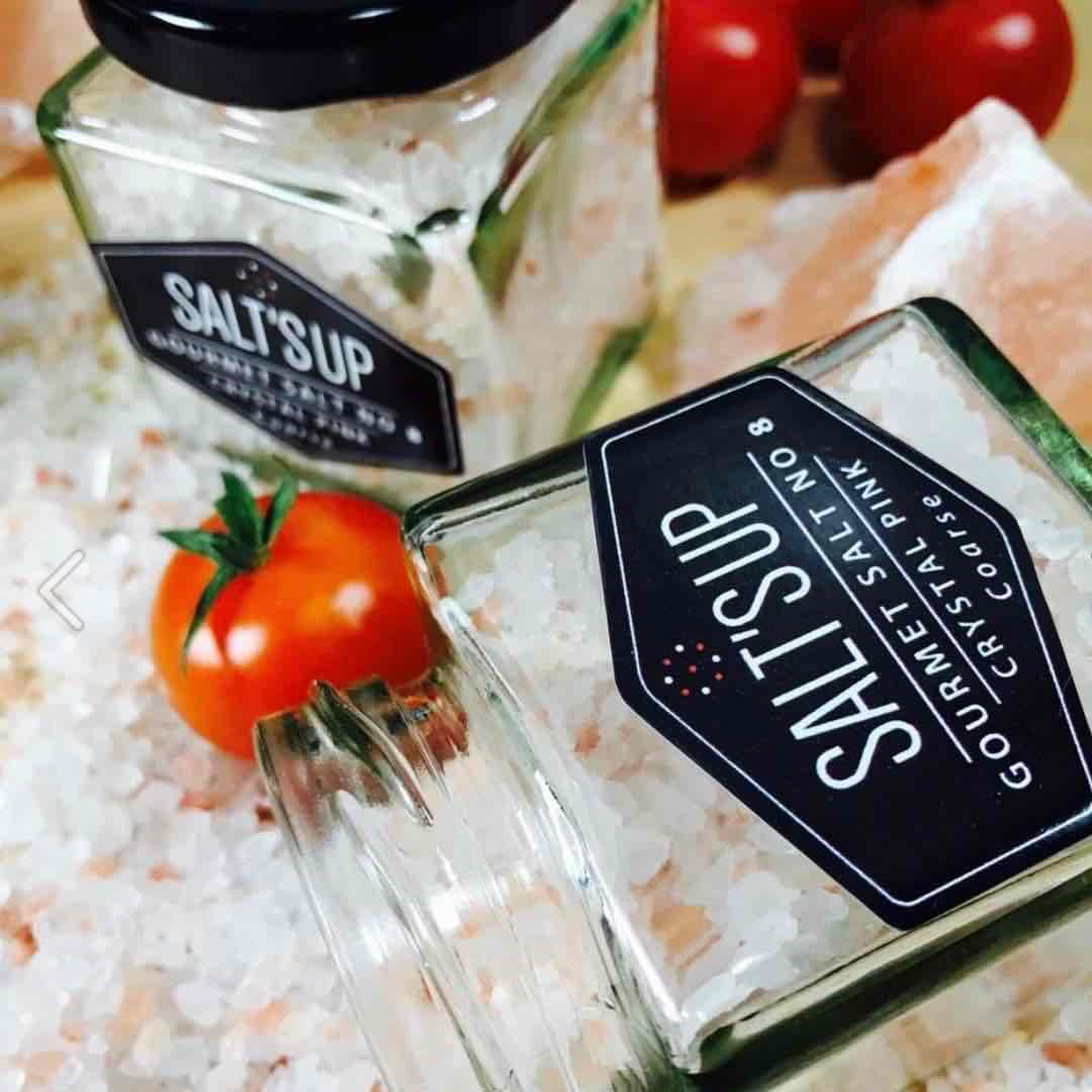 Gourmet coarse pink salt crystals in glass jars, with tomatoes and salt rocks nearby.