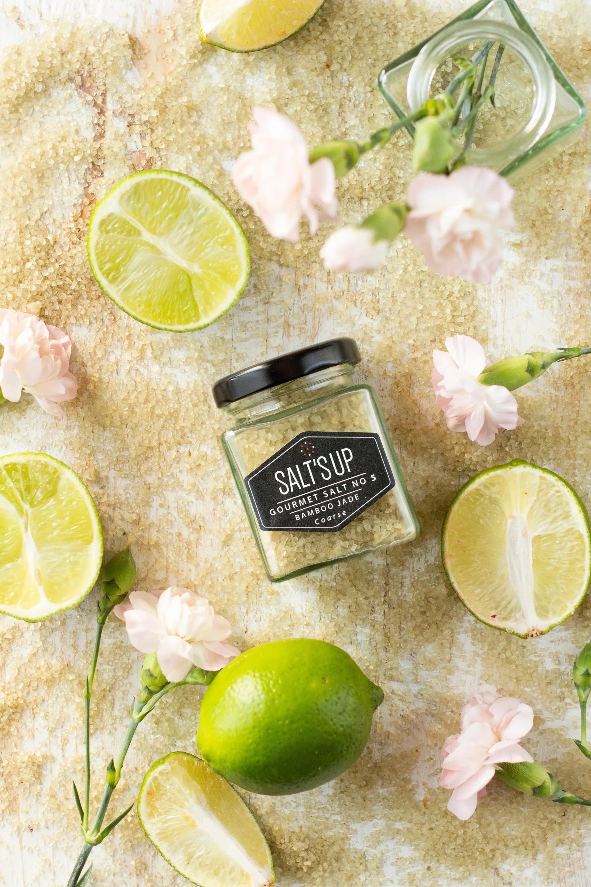 Bamboo Jade gourmet salt jar on coarse salt with lime slices and pink flowers