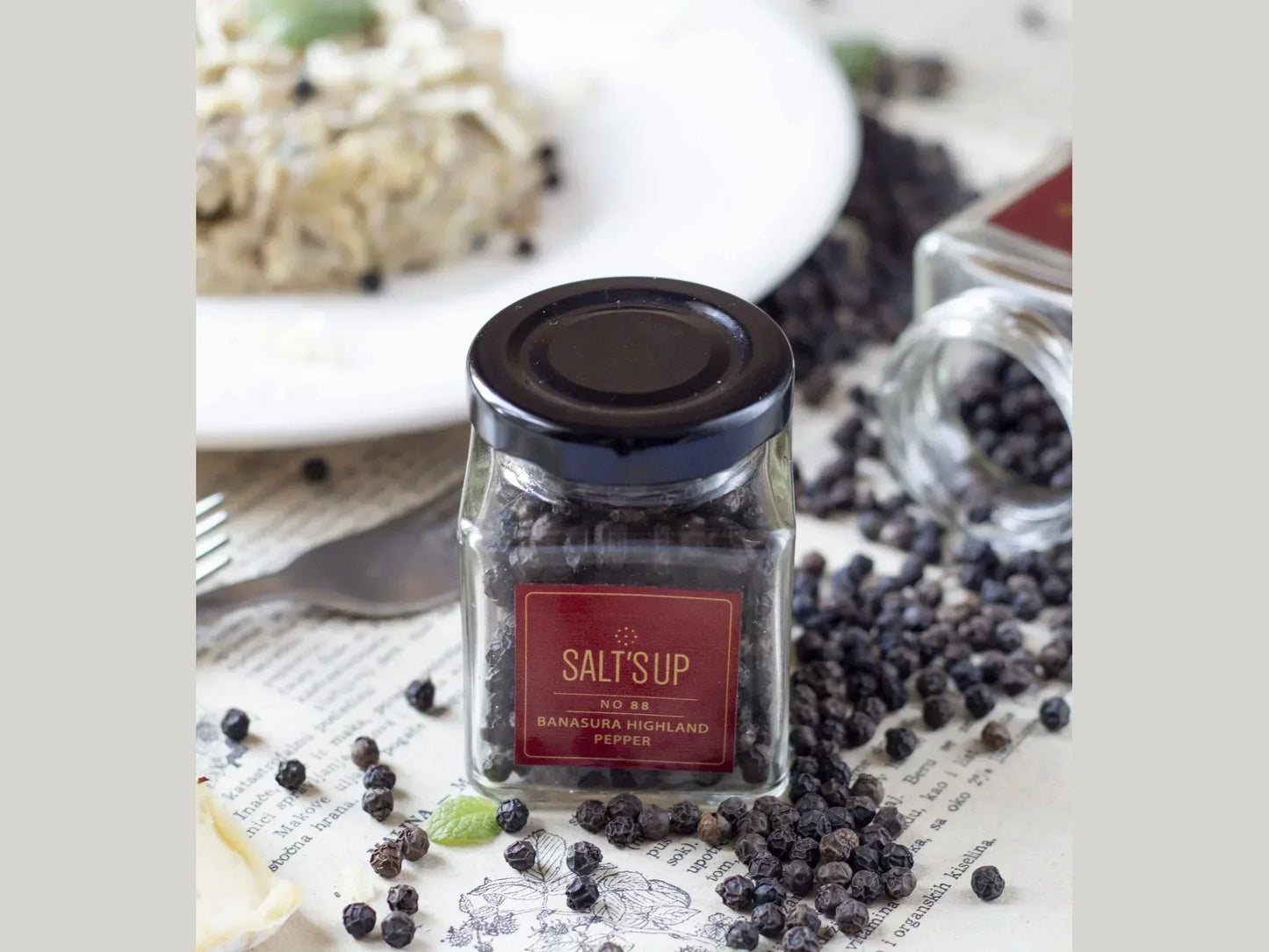 Glass jar of Banasura Highland peppercorns on table with scattered pepper and a food plate