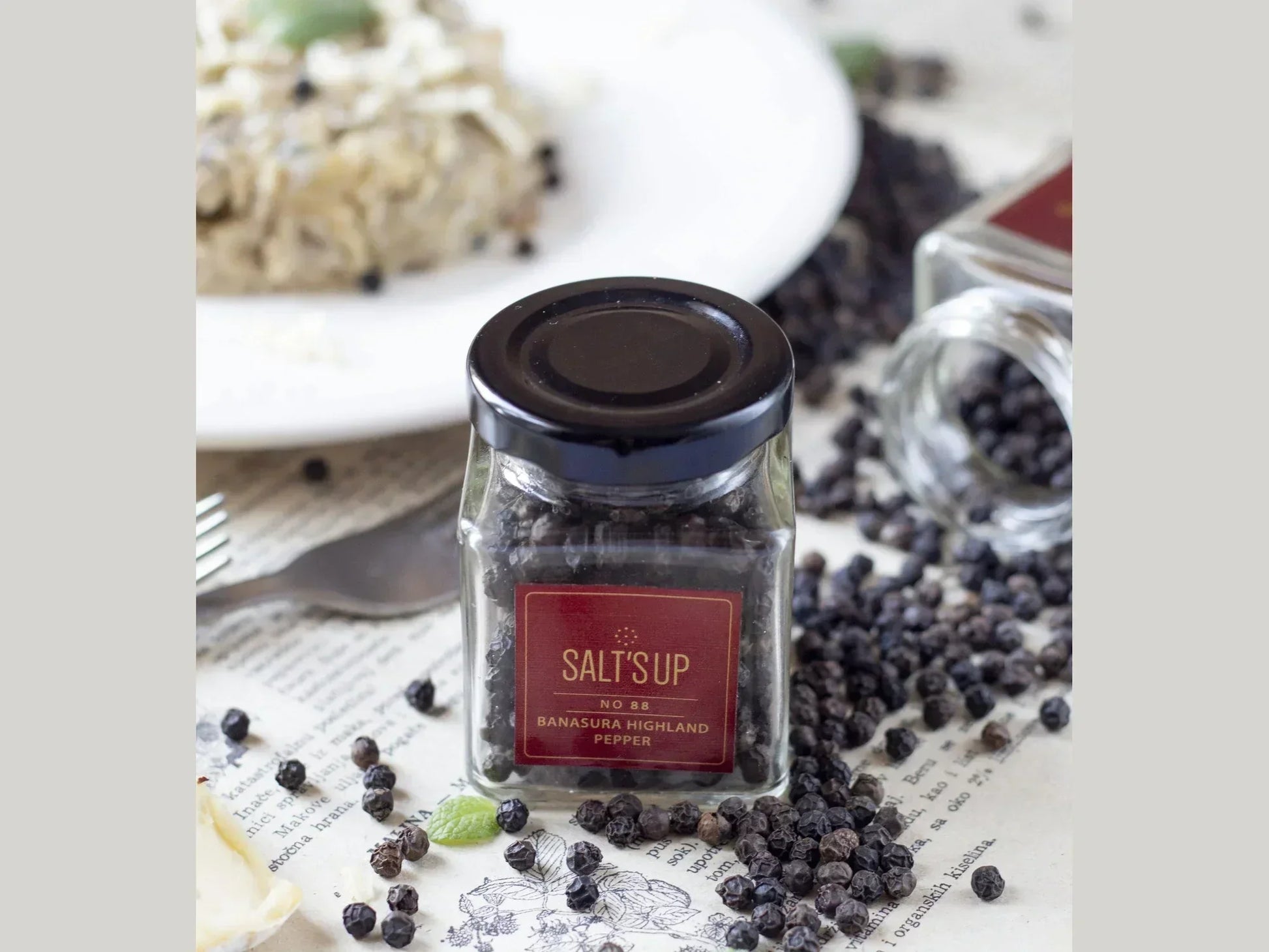 Glass jar of Banasura Highland peppercorns on table with scattered pepper and a food plate