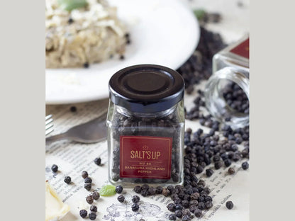 Glass jar of Banasura Highland peppercorns on table with scattered pepper and a food plate