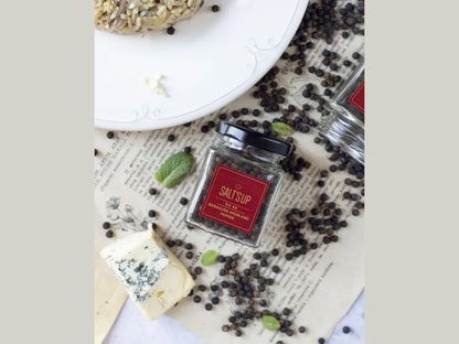 Glass jar of Banasura Highland peppercorns with cheese, herbs, and pepper scattered on paper