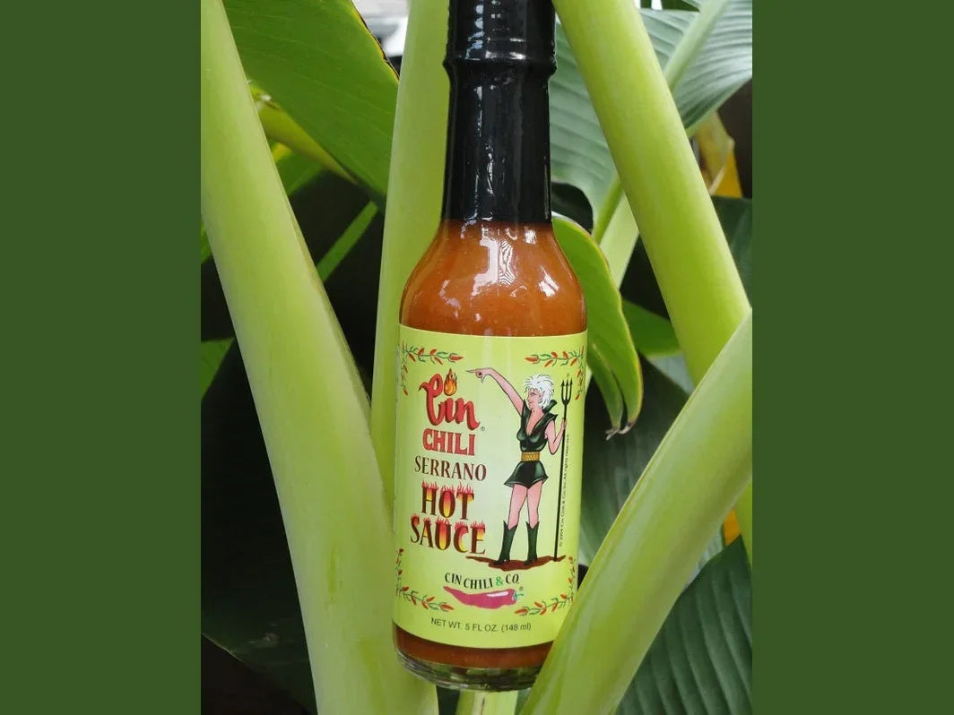 Serrano chili hot sauce bottle with illustrated label displayed among green tropical leaves