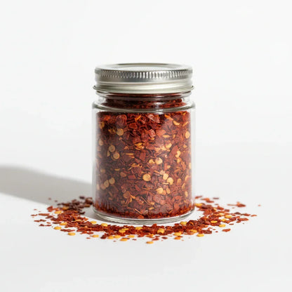 Glass jar of gourmet red chili flakes with metal lid on white background, Spice n Slice Market.