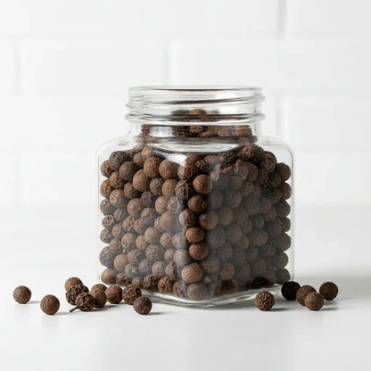 Glass jar filled with whole black peppercorns on white background, some peppercorns scattered