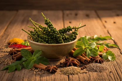 Fresh green peppercorns in a bowl with assorted whole spices and herbs on rustic wood