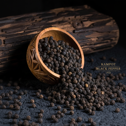 Kampot black pepper PGI peppercorns spilling from carved wooden bowl on dark slate