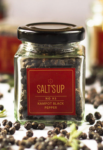 Glass jar of Kampot black peppercorns labeled SALT'S UP, surrounded by loose peppercorns