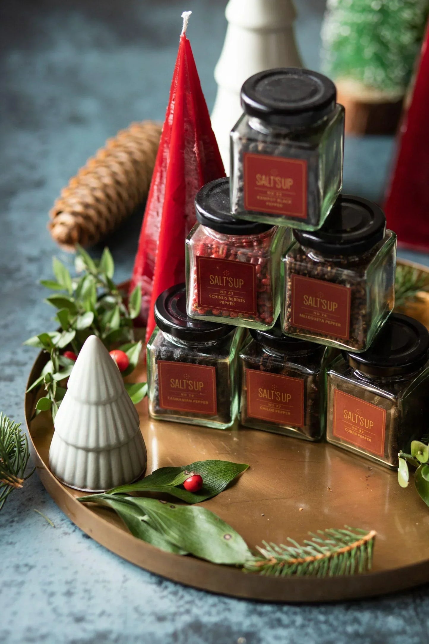 Assorted gourmet pepper jars on a festive tray with candles, pine, and holiday decor.